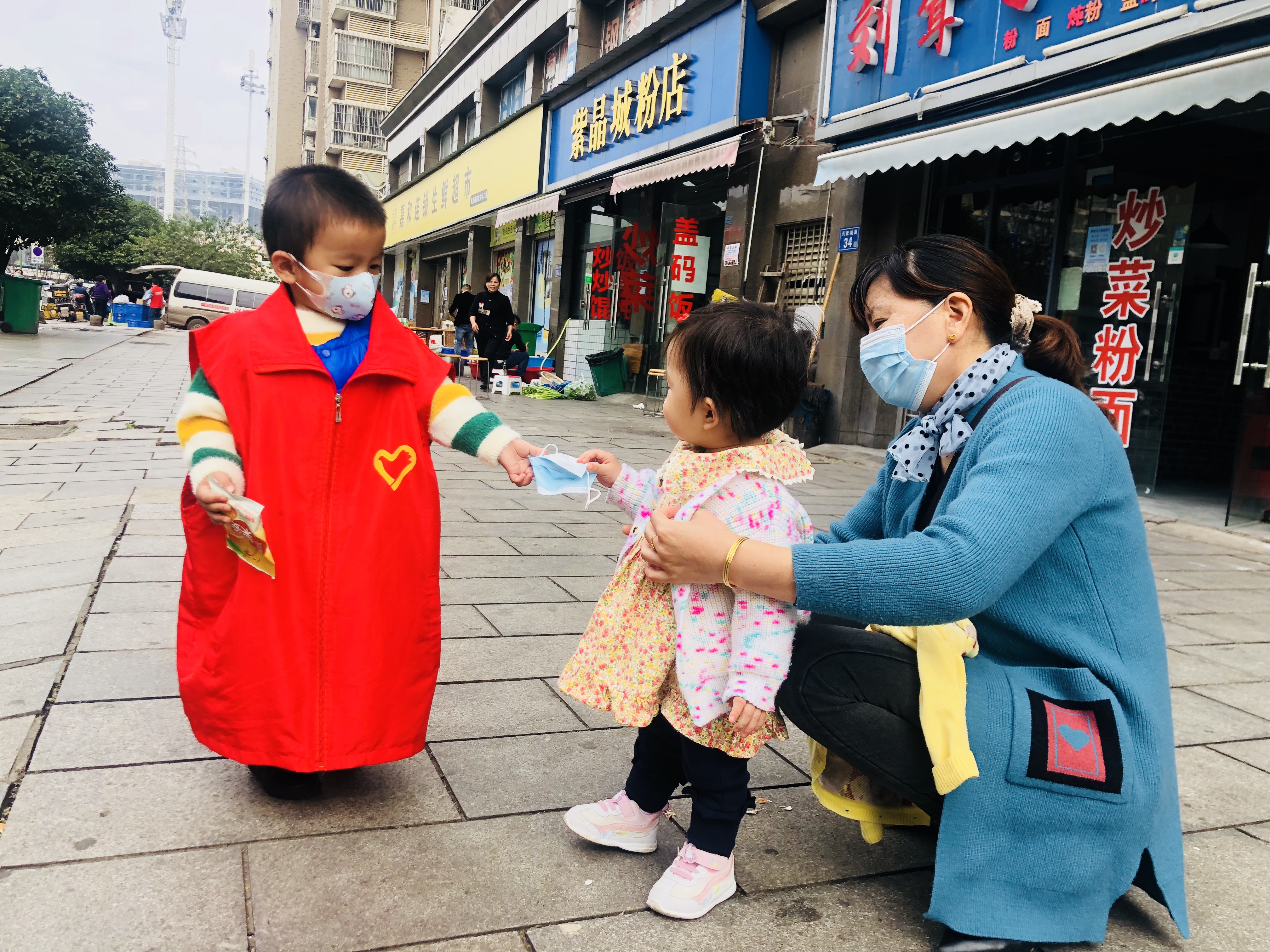 26日,，跟著媽媽來“上班”，3歲萌娃志愿者鄒涵睿給沒佩戴口罩的居民送口罩,。